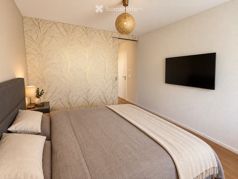 Modern bedroom with a large bed, nightstand with lamp, wall-mounted TV, and a decorative pendant light. The walls feature a stylish, abstract pattern.