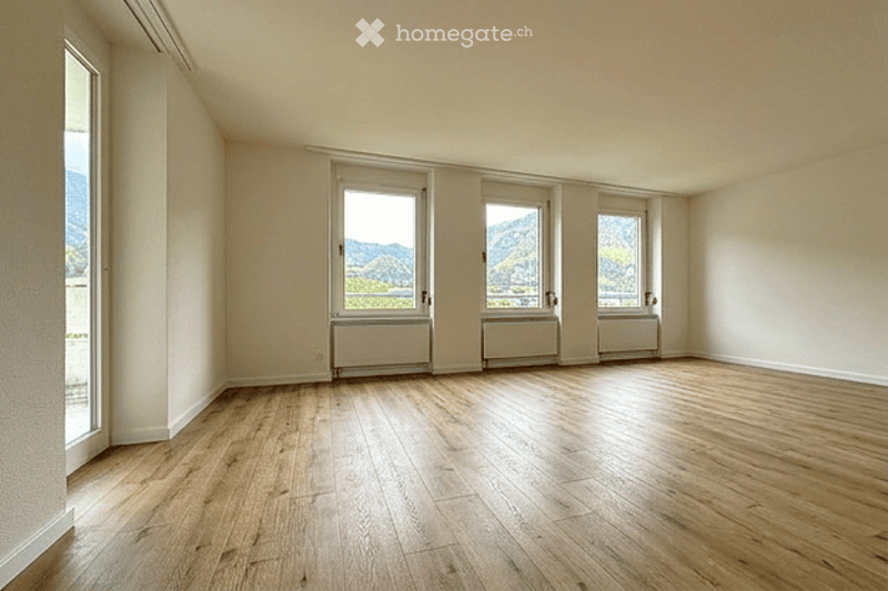 Empty room with wooden flooring, white walls, and large windows offering a scenic mountain view. No furniture or equipment present.
