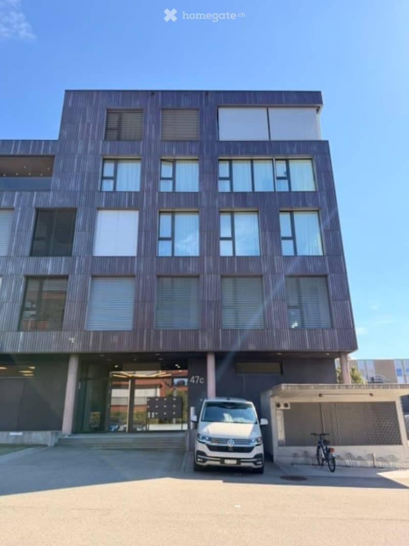 Modern multi-story building with large windows and a covered entrance area. A white van is parked in front, and a bicycle is visible near the entrance.
