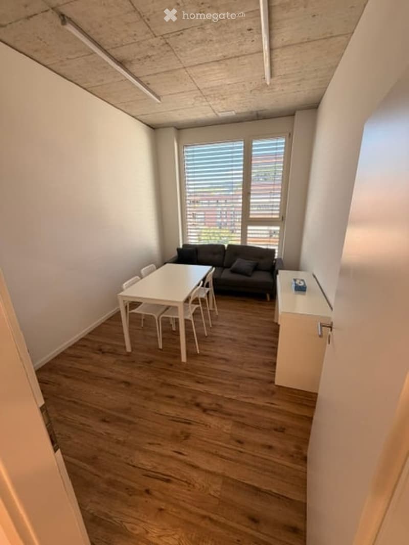 Bright, modern room with wooden flooring, white walls, and large windows. Features a black sofa, white dining table with chairs, and a small cabinet.