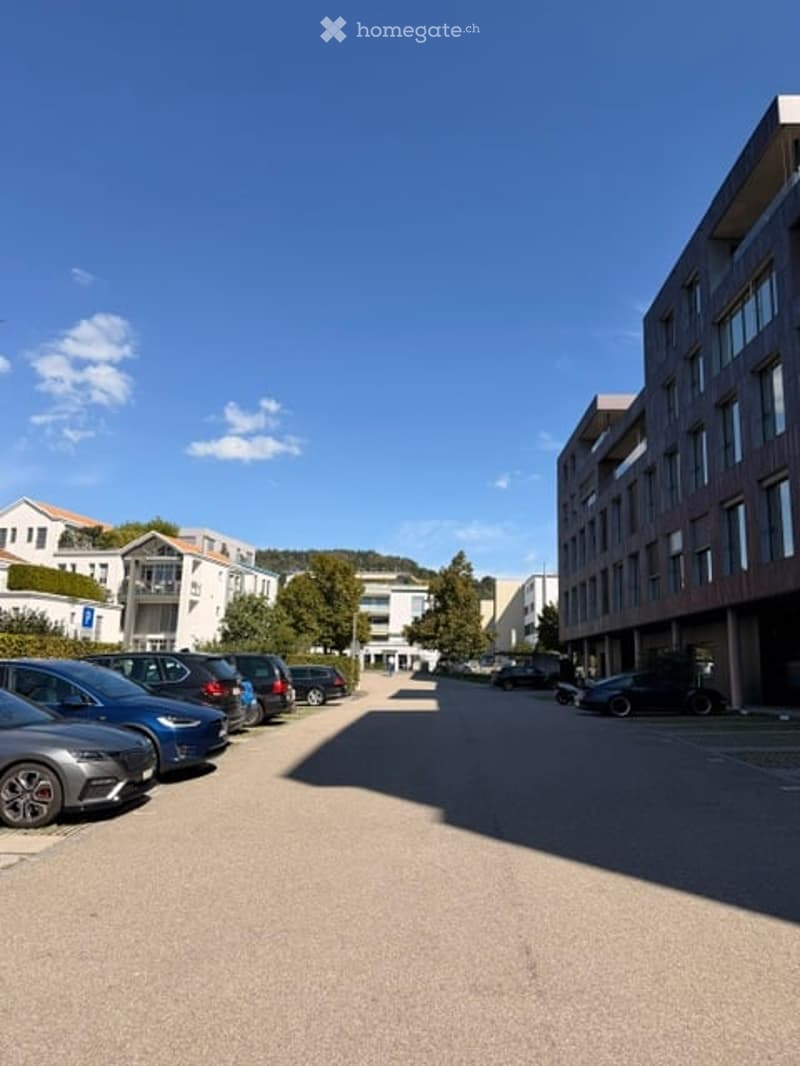 Modern multi-story building with large windows and balconies, surrounded by parked cars and greenery.
