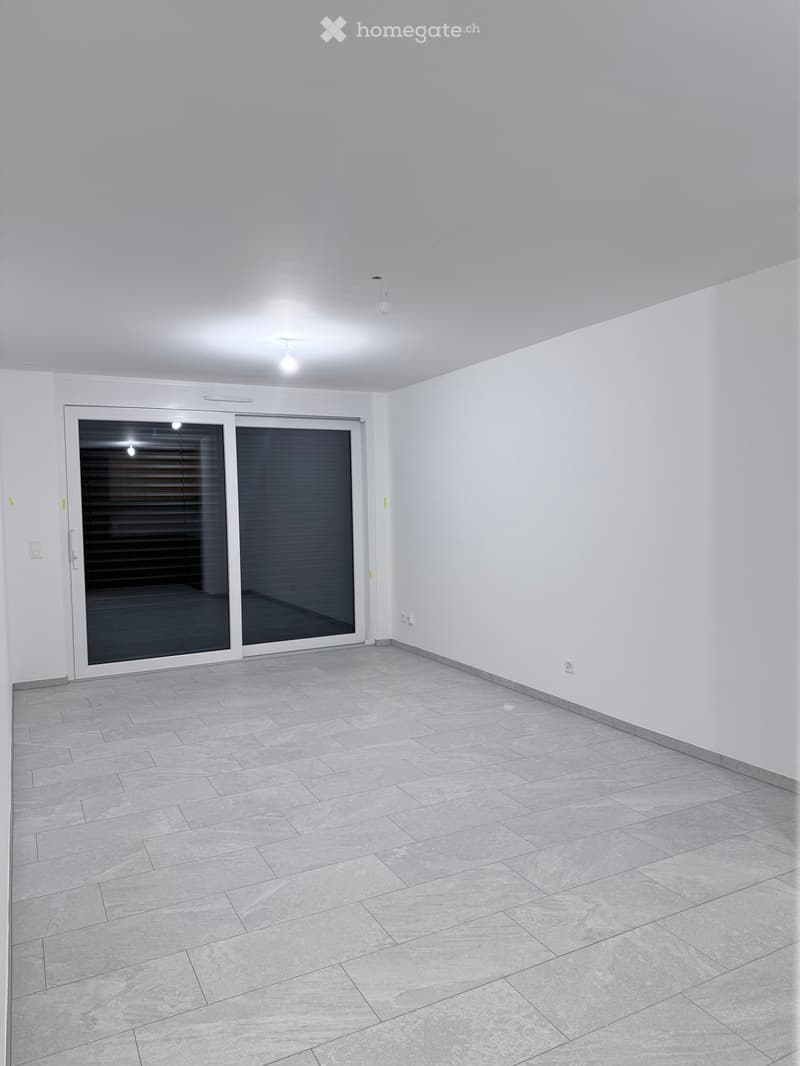 Empty room with white walls, grey tiled floor, and large sliding glass door with blinds.