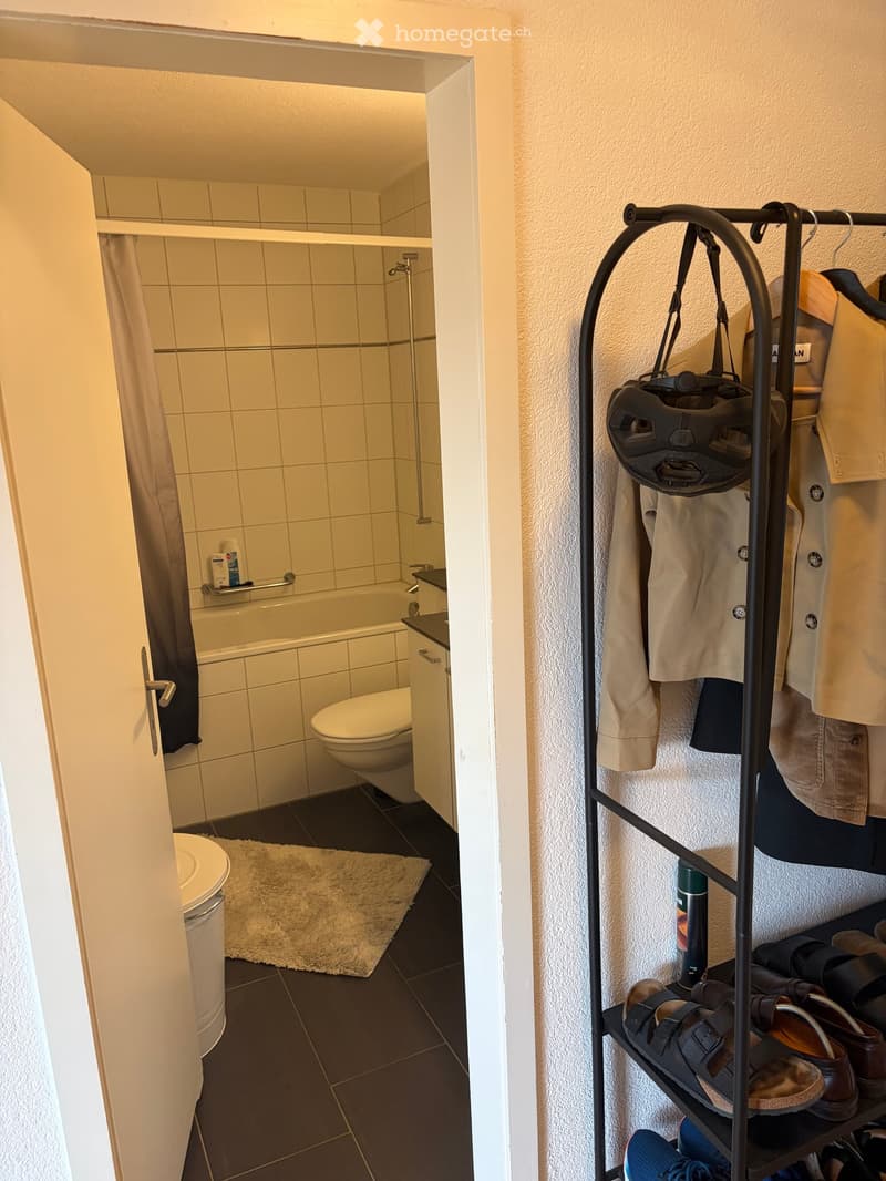 Bathroom with a bathtub, toilet, shower, and a coat rack with clothes and shoes. The floor is tiled, and there is a small rug in front of the bathtub.