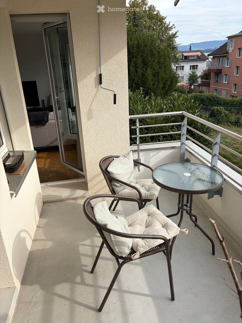 Balcony with two wicker chairs and a small round glass table, overlooking a green area and distant buildings.