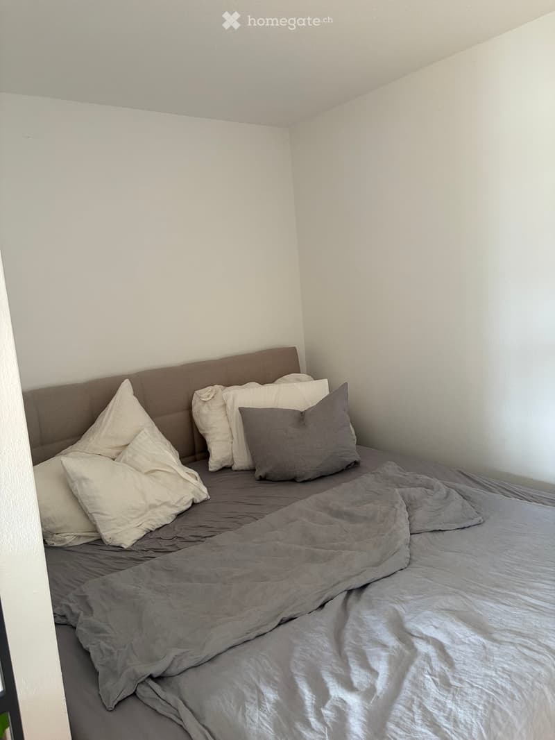 A small bedroom with a neatly made bed featuring a grey duvet and multiple pillows, positioned against a white wall.