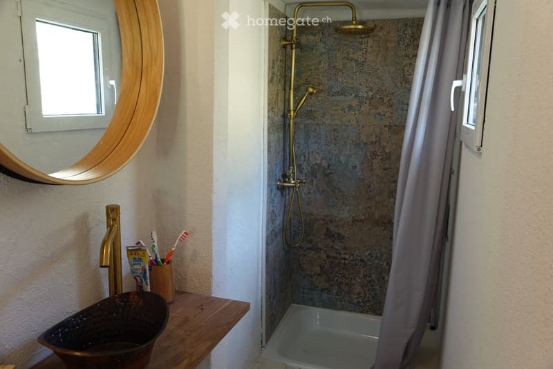 Bathroom with a wooden shelf holding a toothbrush holder, toothpaste, and a dark-colored bowl. A round wooden-framed mirror is mounted on the wall, and a shower with a gold-colored showerhead is visible in the background.