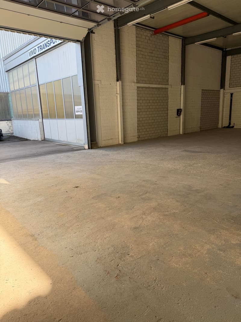 Empty garage with concrete floor, brick walls, and a partially open garage door leading to a building with glass windows and signage reading "VIVO TRANSPORT".