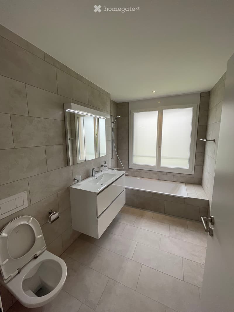 Modern bathroom with a toilet, sink, bathtub, and large window providing natural light. Features a sleek vanity with a mirror and modern fixtures.