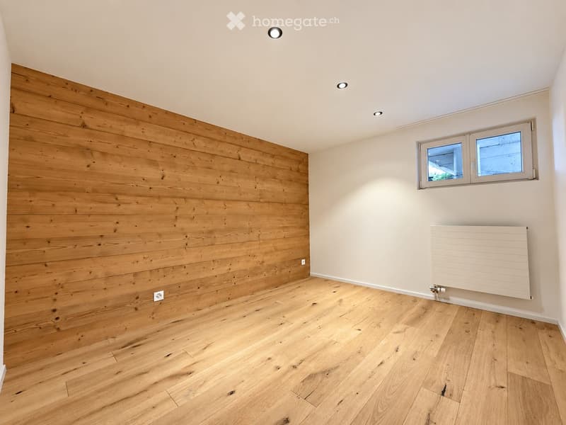 Empty room with wooden flooring and a wooden accent wall, featuring a window and a radiator.