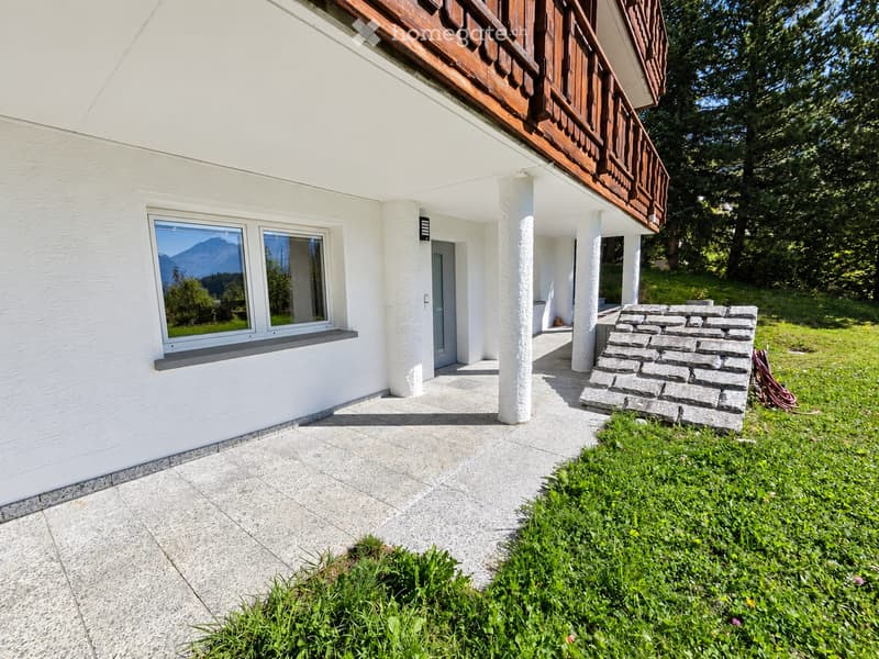 A modern white house with a wooden balcony, large windows, and a stone pathway leading to the entrance, surrounded by greenery and a scenic mountain view.
