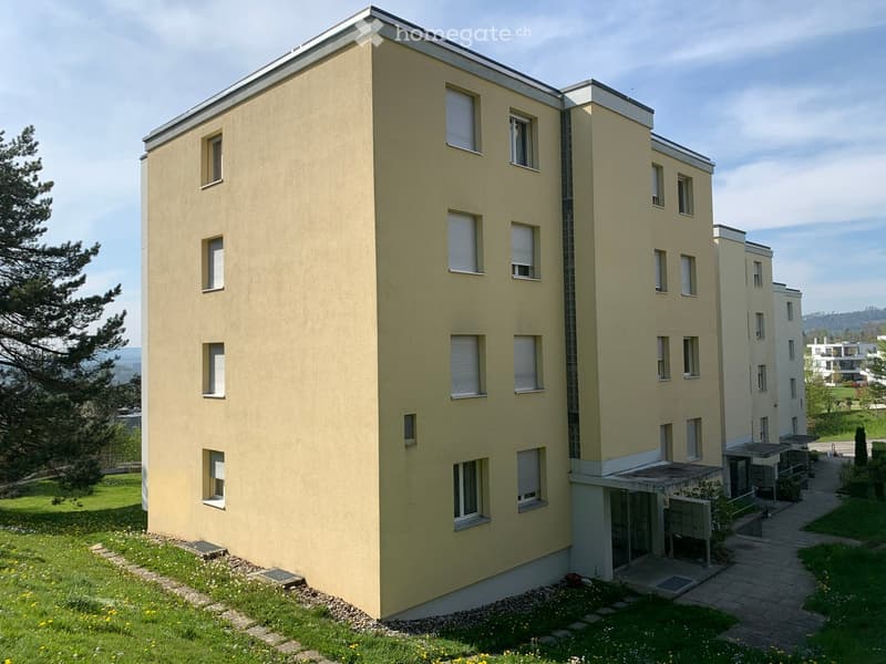 Modern multi-story building with light yellow facade, multiple windows with white frames, and a landscaped garden area with pathways and small trees.