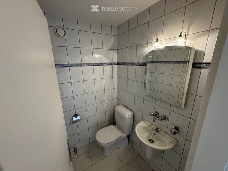 Small bathroom with tiled walls and floor, featuring a toilet, sink, mirror, and wall-mounted light fixture.