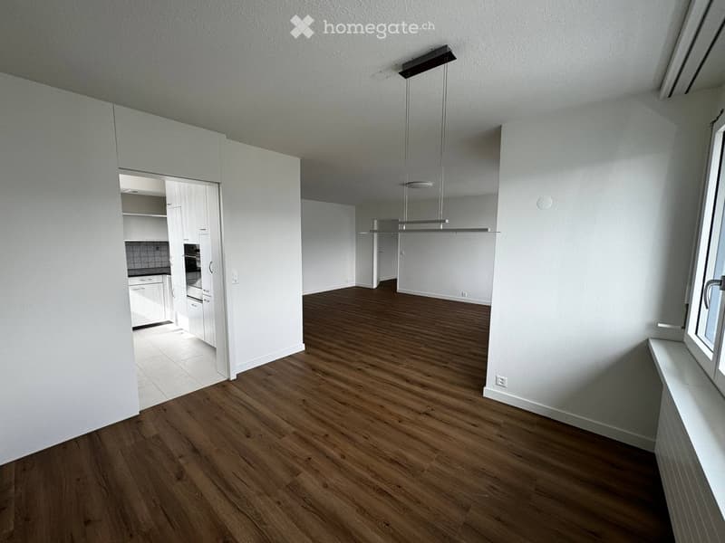 Empty room with wooden flooring, white walls, and a modern pendant light fixture. The room is connected to a small kitchen area with white cabinets and a tiled backsplash.
