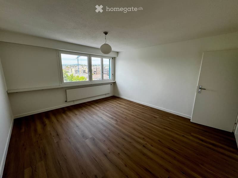 Empty room with wooden flooring, white walls, a window with a view, a radiator, and a single light fixture on the ceiling.