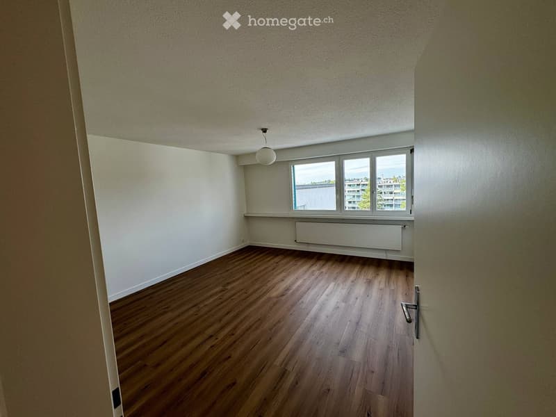 Empty room with wooden flooring, white walls, and a large window offering a view of buildings and greenery outside.