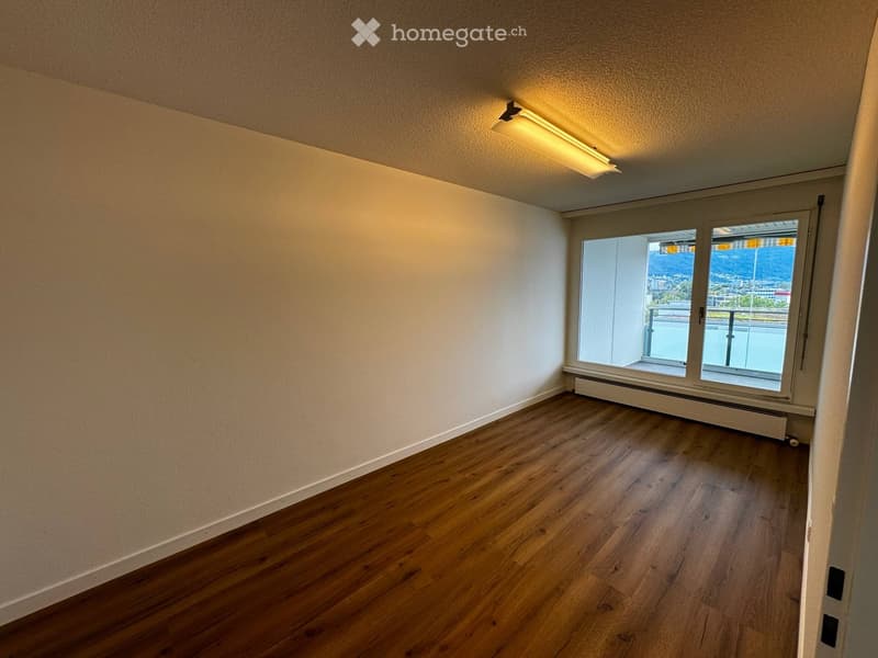 Empty room with wooden flooring, white walls, and large windows offering a view of the city and mountains.