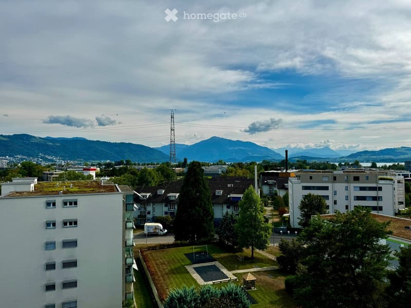 A view of a residential area with multiple apartment buildings, green spaces, and a playground, with mountains in the background.