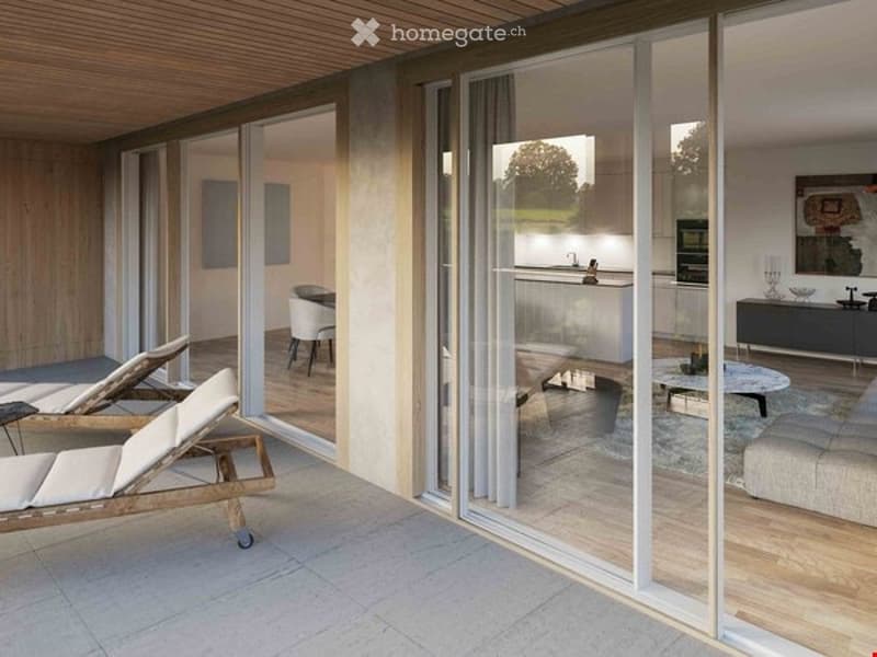 Modern, well-lit living space with large glass sliding doors connecting to a patio. The living area features a white sofa, coffee table, and a dining area with chairs and a table. The kitchen area includes cabinets, a faucet, and an oven. A mirror and an