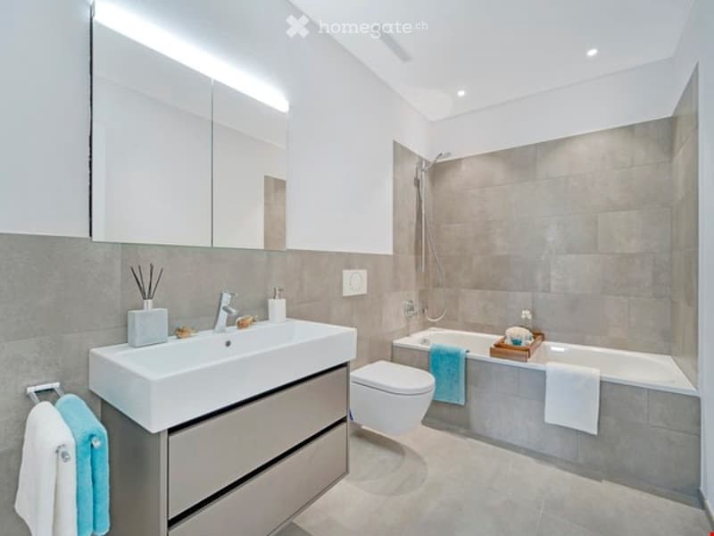 Modern bathroom with a white sink, chrome faucet, large mirror, toilet, bathtub, and towels neatly placed on the edge of the tub.