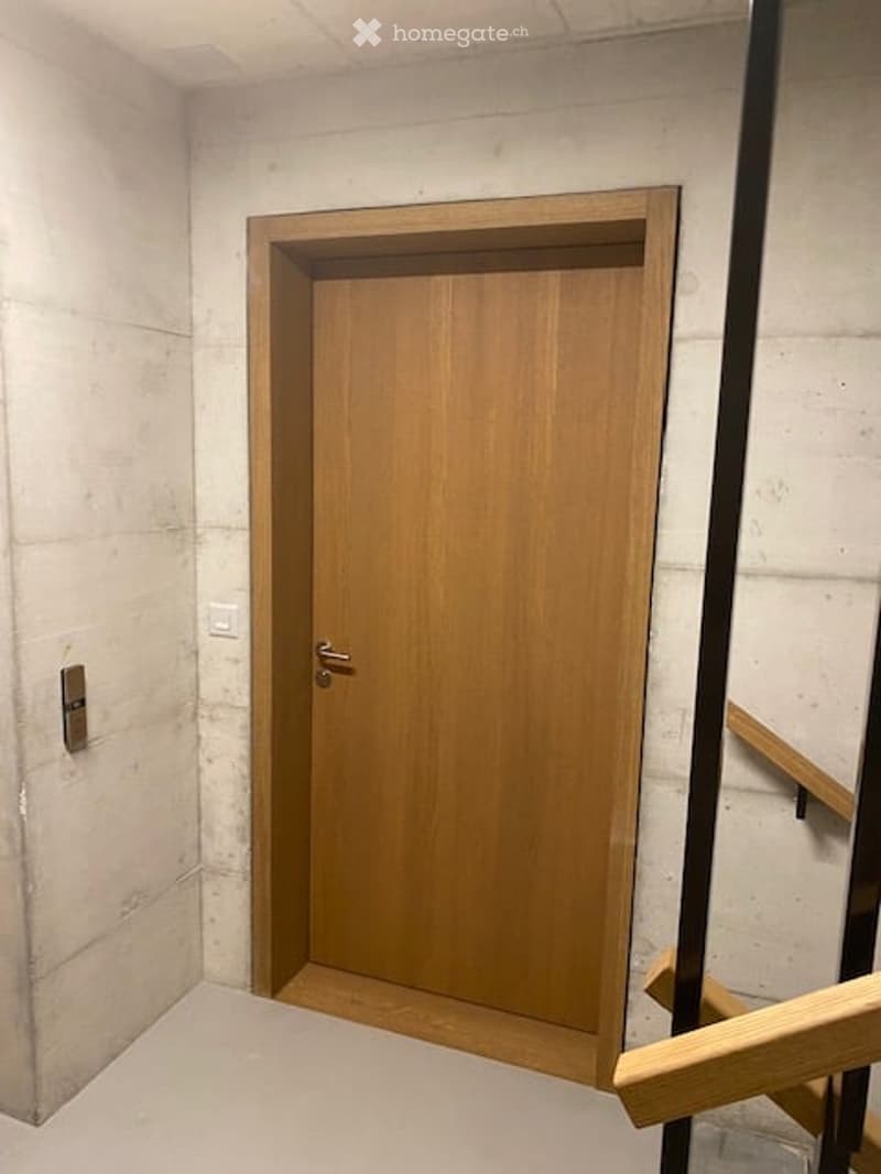 A narrow hallway with a wooden door and a staircase railing visible. The walls are concrete, and the floor is tiled.