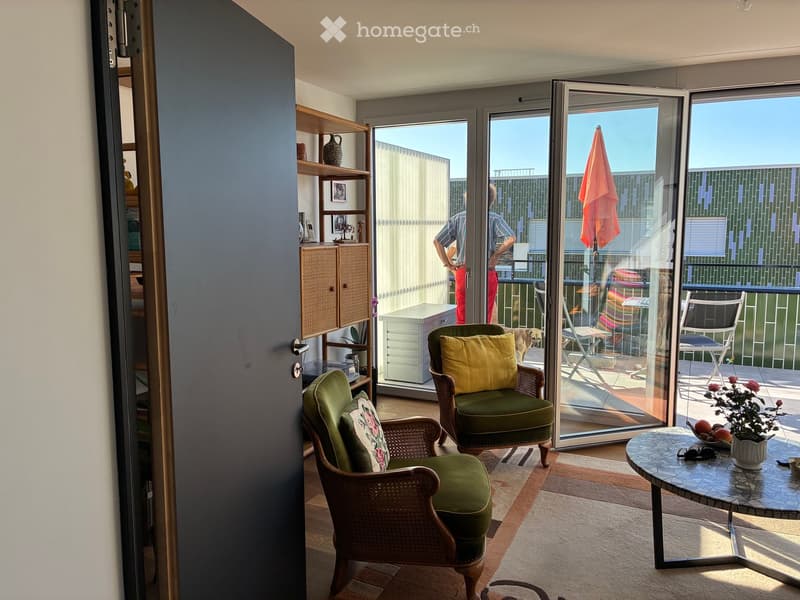 Bright living space with large glass doors opening to a balcony, featuring wicker chairs, a round table, shelves, and a cabinet.