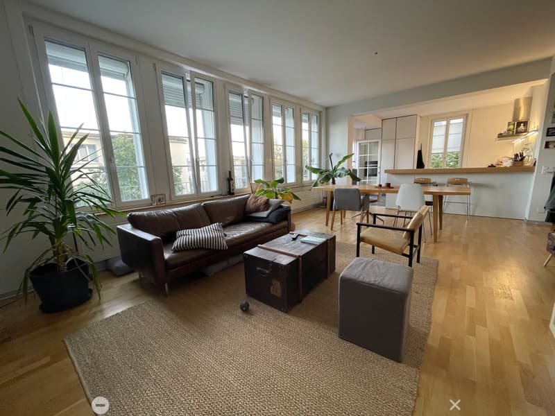 Modern living room with large windows, leather sofa, coffee table, ottoman, dining area with table and chairs, and plants.