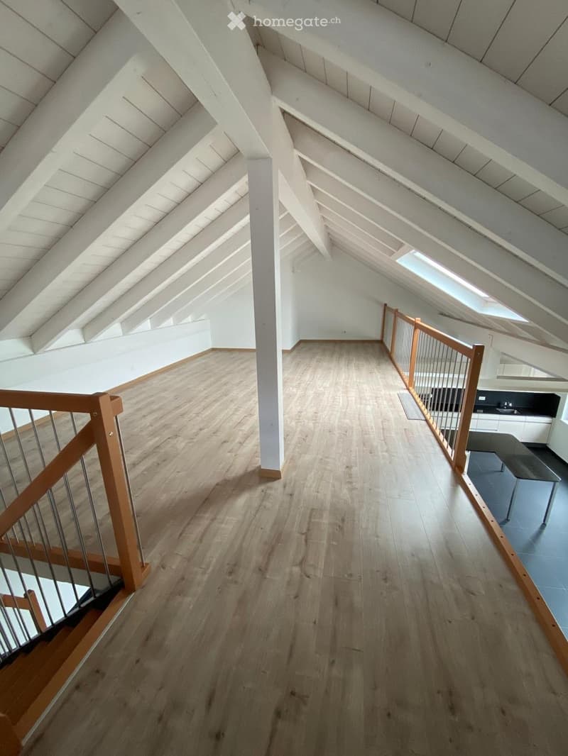Bright, open hallway with wooden flooring, white walls, and exposed wooden beams on the ceiling. Features a wooden railing and a small kitchen area visible at the end.