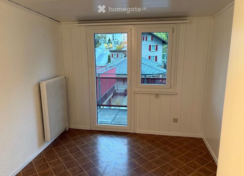 Empty room with tiled floor, white walls, radiator, and large window with a view of the street and neighboring buildings.