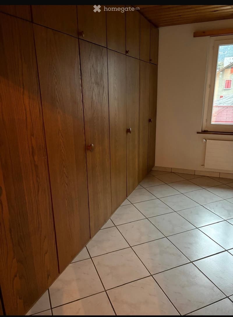 Empty room with tiled floor and wooden built-in closet, featuring a window with a view of a neighboring building.