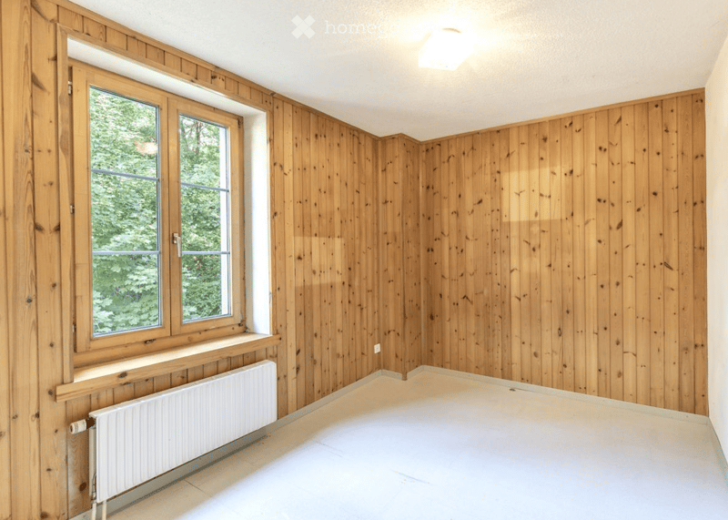 Empty room with wooden panel walls, large windows, and a radiator. No furniture or equipment present.