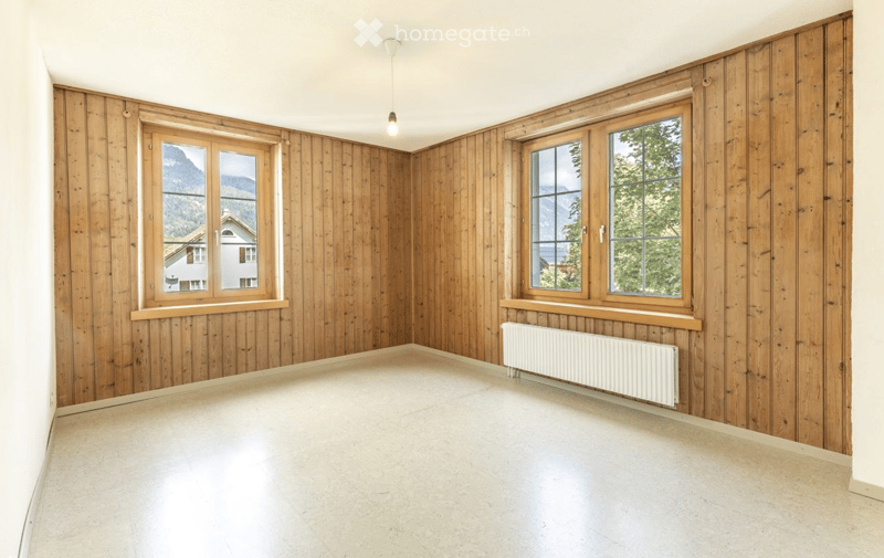 Empty room with wooden panel walls, large windows, and a radiator. The space is unfurnished with a clean, minimalist design.