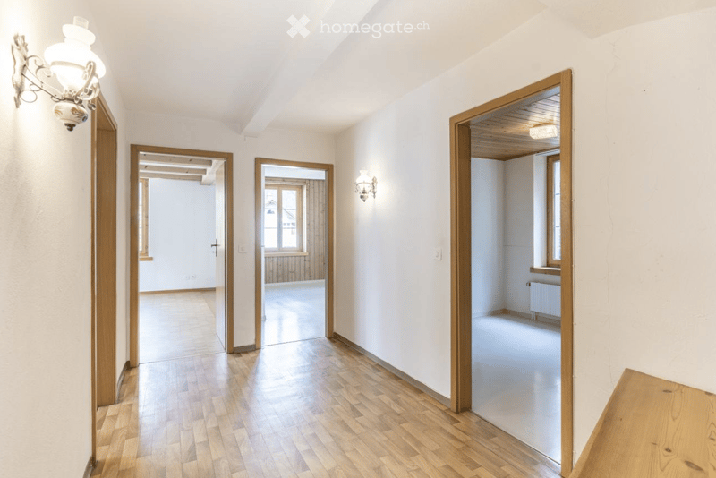 Empty hallway with wooden flooring, white walls, and several open doors leading to other rooms. Light fixtures are mounted on the walls.