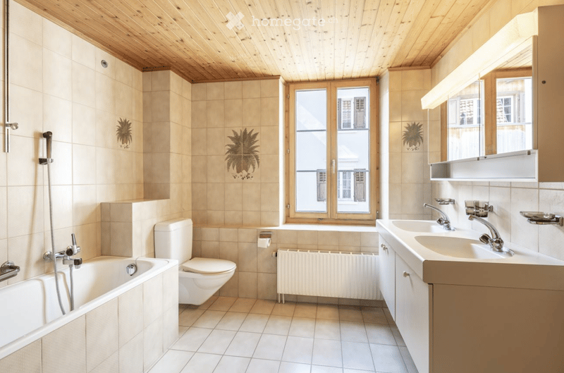 Bright bathroom with beige tiles, wooden ceiling, bathtub, toilet, double sink, and large window offering a view of the street.