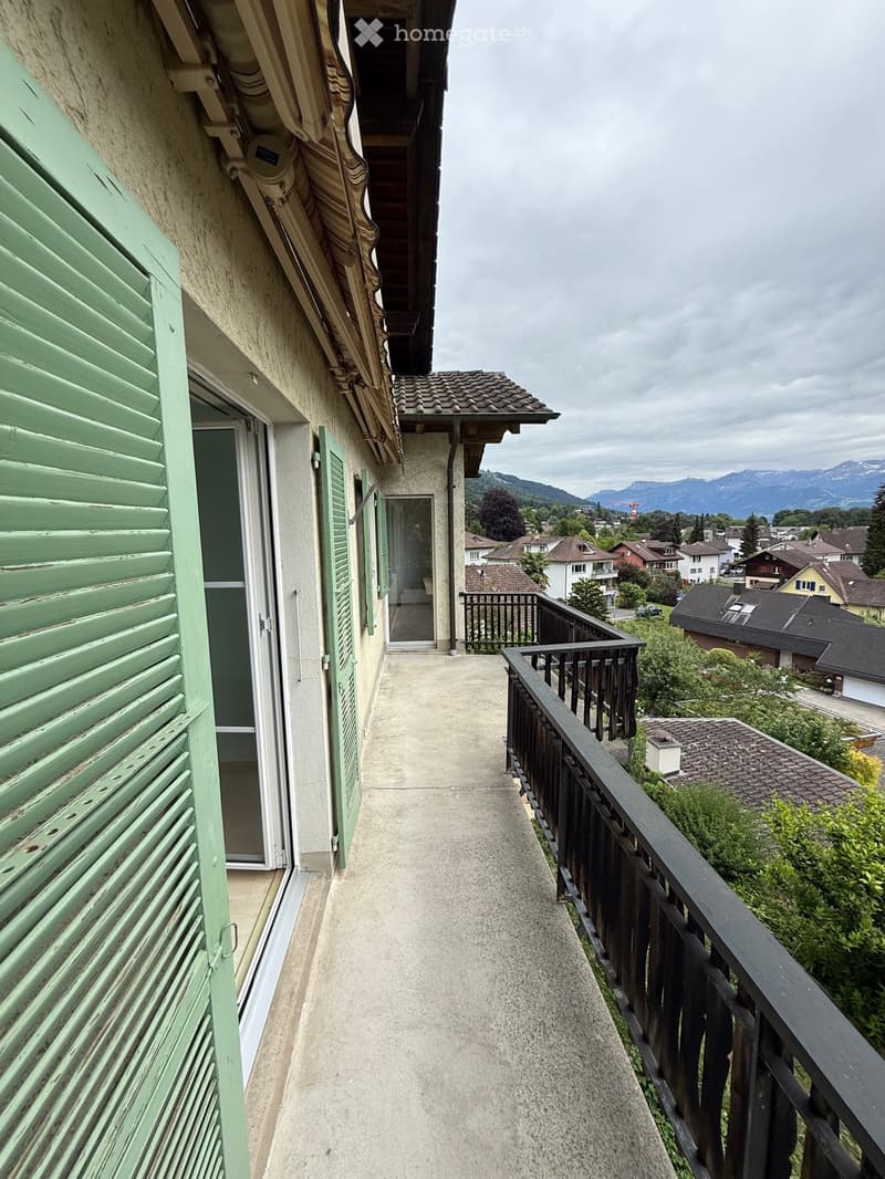 Balcony with a view of a town and mountains, featuring a concrete floor, black railing, and green shutters on the adjacent building.
