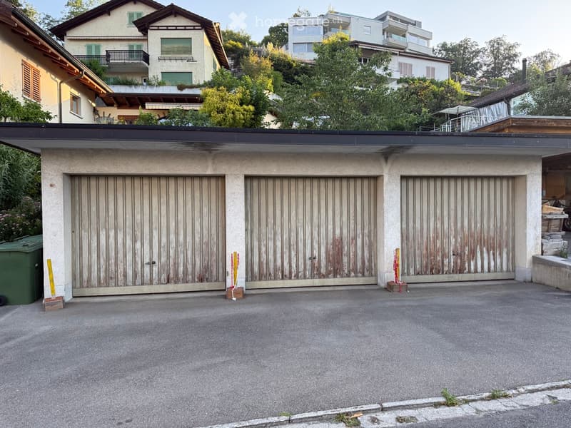 A multi-car garage with three large wooden garage doors, situated in a residential area with neighboring houses and greenery visible in the background.