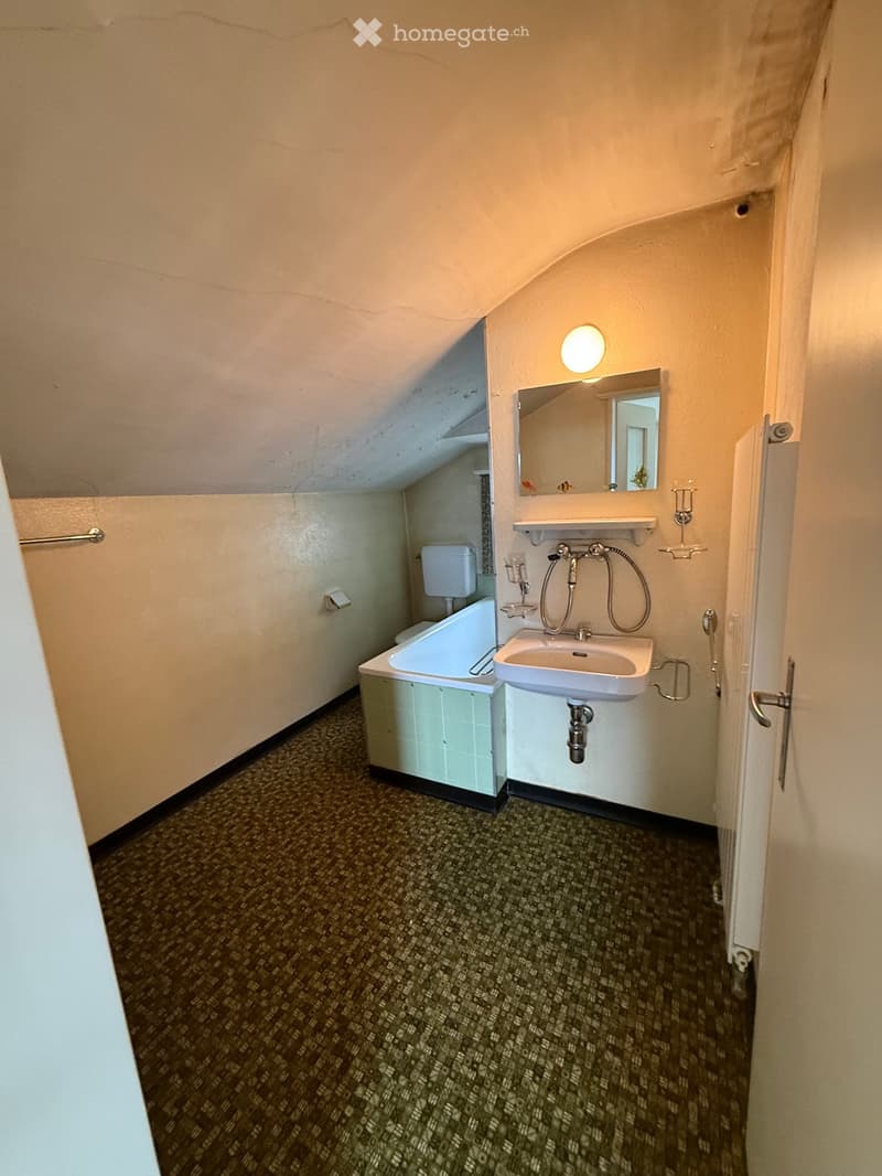 Small bathroom with a sink, bathtub, toilet, mirror, and light fixture. The room has a slanted ceiling and patterned carpet flooring.