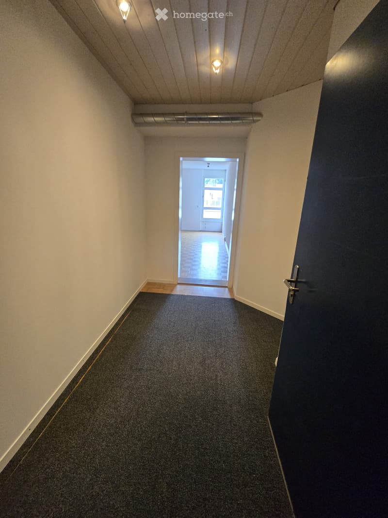 A narrow, carpeted hallway with light-colored walls, a wooden baseboard, and a closed door on the right. Natural light streams in from an open doorway at the end, revealing a glimpse of another room with a window and a tiled floor.