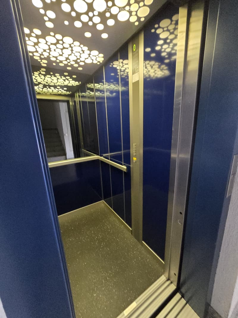 Modern hallway with dark blue walls, reflective surfaces, and a patterned ceiling with circular light fixtures. The floor is a polished, metallic-looking material.
