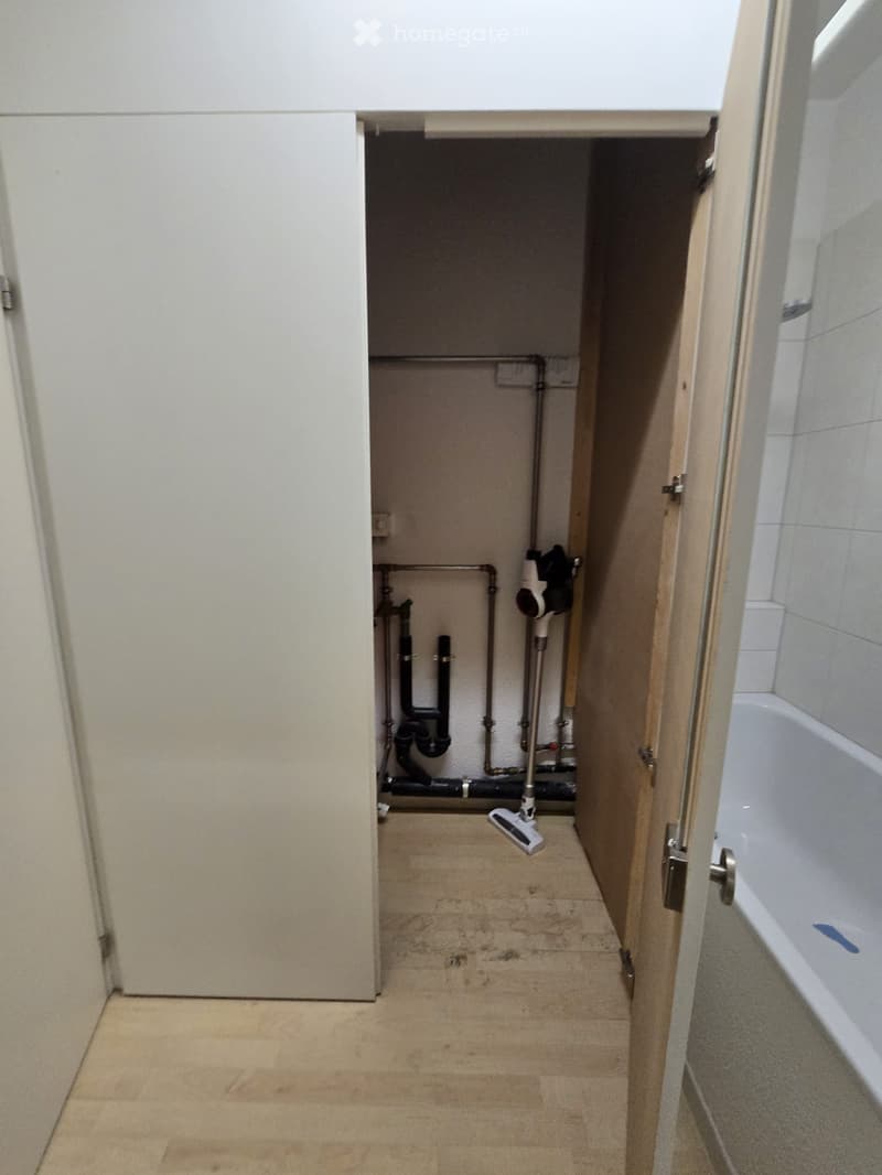 Empty room with exposed pipes and a vacuum cleaner, adjacent to a tiled bathroom with a bathtub.