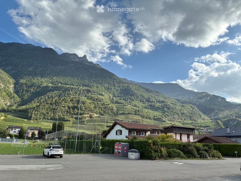 A small house with a red-tiled roof and white walls, situated in a scenic valley with green hills and mountains in the background. A white car is parked in the foreground, and a fenced sports field is visible to the left.