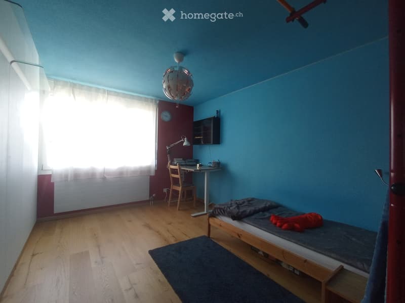 Cozy bedroom with wooden floor, blue walls, a single bed with a dark blue duvet and red pillow, a small desk with a chair, a wall-mounted lamp, and a window with white curtains.