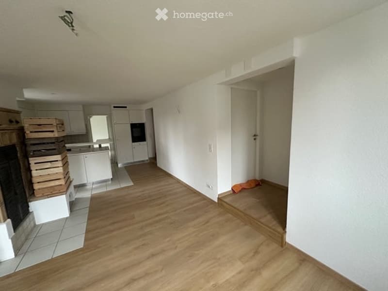 Empty room with wooden flooring, white walls, and a fireplace on the left side. There is a doorway leading to another room with white cabinets and appliances visible.