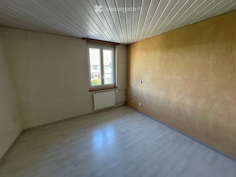 Empty room with wooden flooring, beige walls, a window with a radiator below, and a textured wall on one side.
