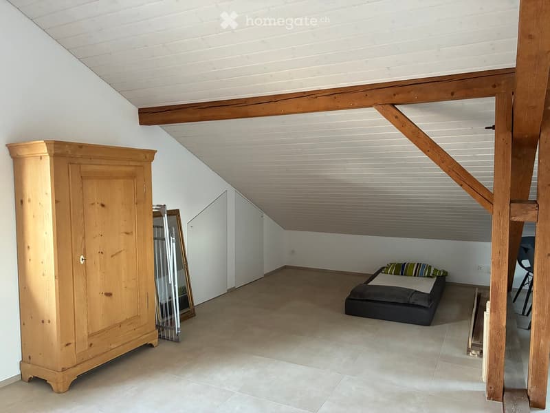 Loft-style bedroom with exposed wooden beams, a large wooden wardrobe, a bed with striped pillows, and a mirror leaning against the wall.
