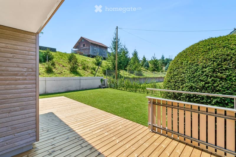 Wooden deck with metal railing, green lawn, and distant view of a wooden house on a hillside.