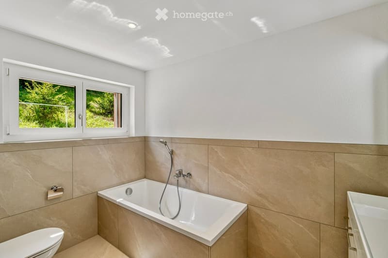 Modern bathroom with a bathtub, toilet, and sink, featuring beige tiles and a large window offering a view of greenery.