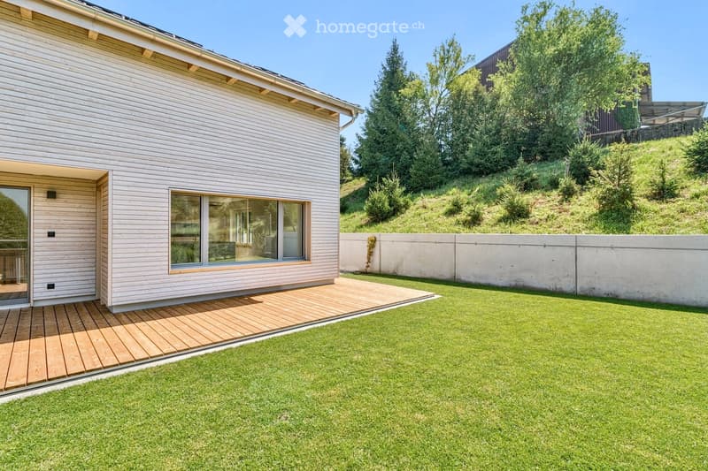 A modern house with a raised wooden deck, large window, and well-maintained lawn surrounded by trees and a concrete wall.