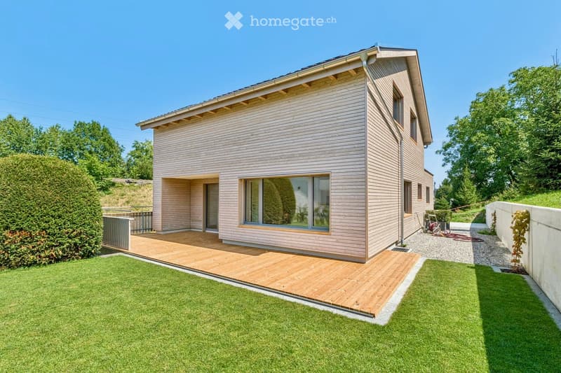 Modern single-story house with light wood siding, large windows, and a wooden deck extending from the entrance. The surrounding area features a well-maintained lawn and a raised wooden deck.