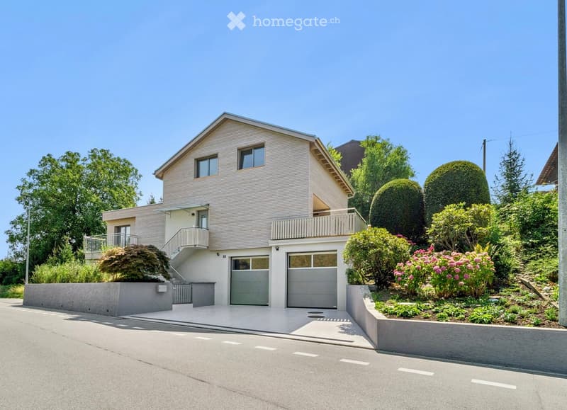 Two-story house with beige siding, grey garage doors, and a balcony on the upper level. The house is surrounded by well-maintained gardens and trees.