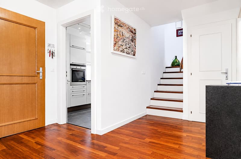 Bright hallway with wooden flooring, white walls, a staircase leading upwards, a framed artwork, and a glimpse of a modern kitchen area with white cabinets and stainless steel appliances.
